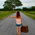 Red-haired woman hitchhiking with vintage suitcase on scenic rural Estonian road. Summer adventure.